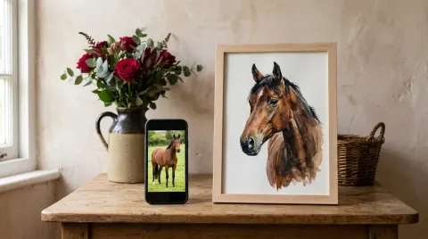 A phone propped on a table showing a photograph of a bay horse in a field, and beside it a natural wood frame holding a watercolour of the same horse.