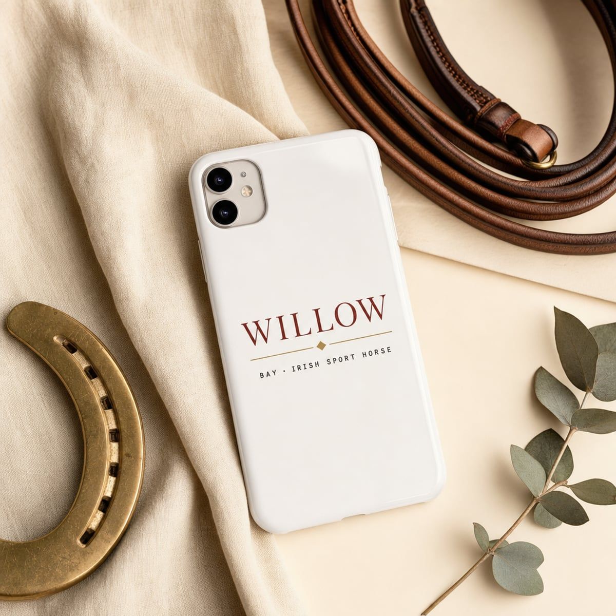 A glossy white iPhone case flat-lay on cream linen showing the printed 'WILLOW' design, beside a brass horseshoe and a sprig of eucalyptus.