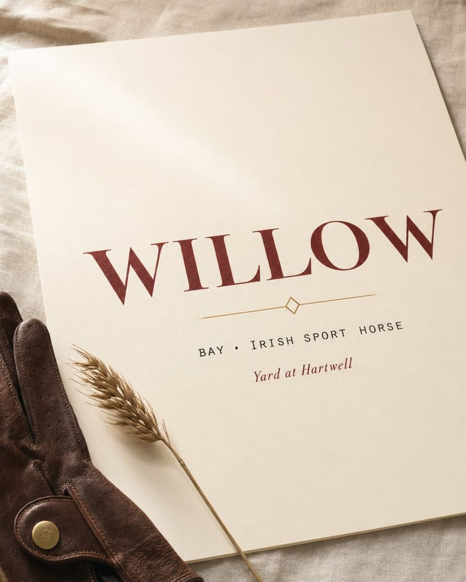 An A3 cream art print propped on cream linen showing 'WILLOW' in oxblood serif capitals, with a leather riding glove and dried wheat at the corner.