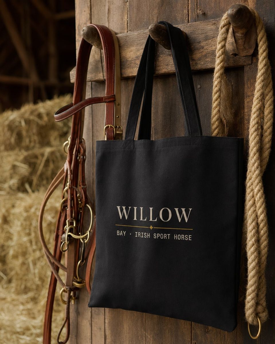 A black cotton tote bag hanging from a barn peg showing the cream-printed design 'WILLOW · BAY · IRISH SPORT HORSE', next to a tan leather bridle and coil of rope.