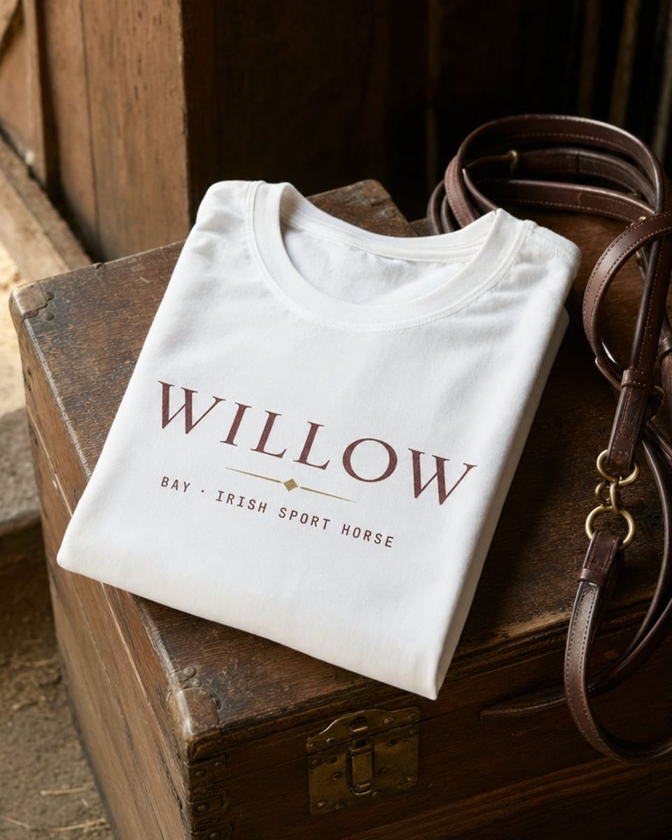 A neatly folded white cotton t-shirt on a wooden stable trunk showing the chest print 'WILLOW · BAY · IRISH SPORT HORSE', next to a coiled leather bridle.